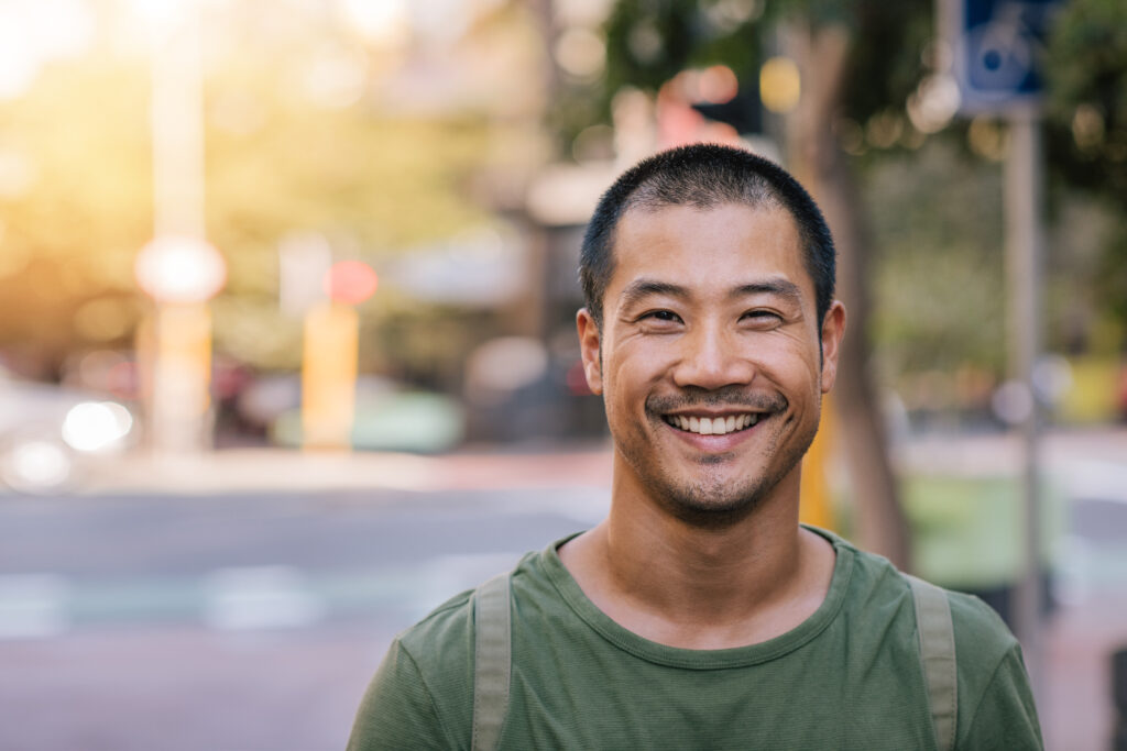 Portrait of a casually dressed handsome young Asian man smiling while standing alone outside on a city street on a sunny day
